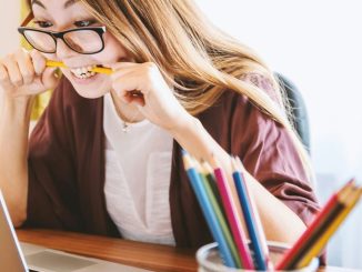 woman biting pencil while sitting on chair in front of computer during daytime