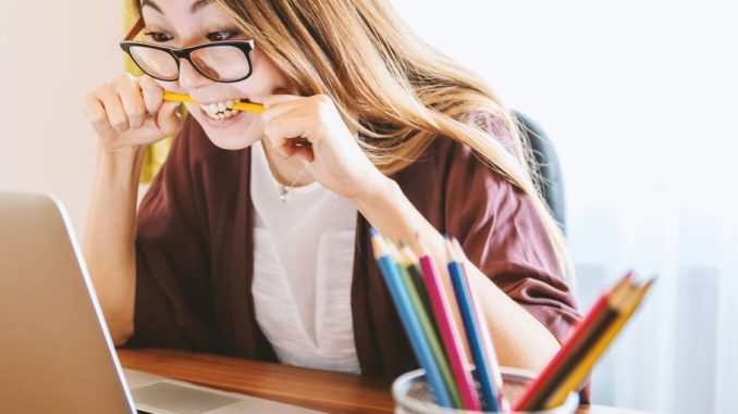 woman biting pencil while sitting on chair in front of computer during daytime